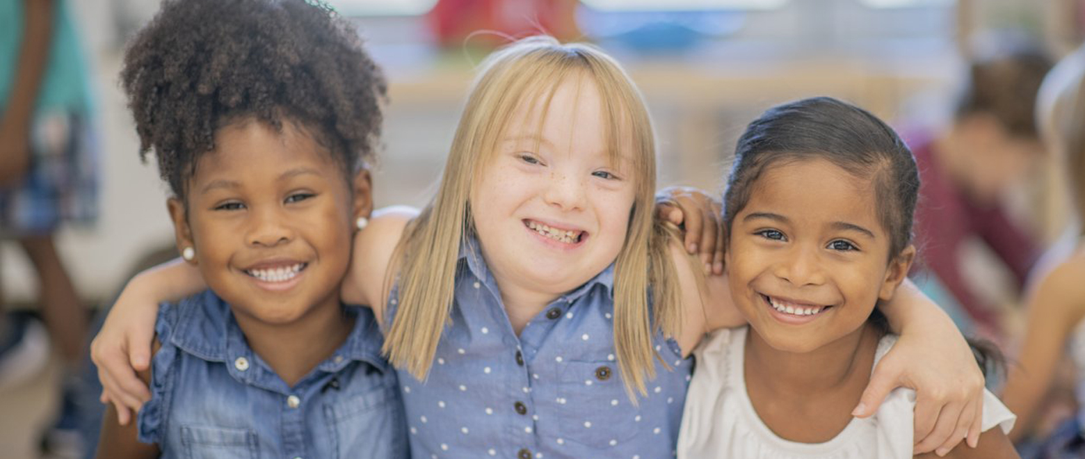 Três meninas se abraçando e sorrindo sendo que a do meio possui Síndrome de Down